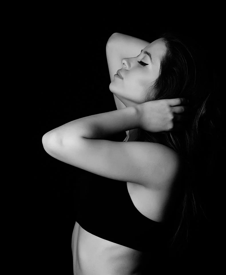 A black and white close-up of a woman in a three-quarter view, eyes closed and serene. Her arms drape over her head, highlighting her shoulders. Long hair flows down her back. Strong shadows and a black background draw focus to her, with a dark sports bra visible. - Diagnostic Laparoscopy in Coral Gables, FL