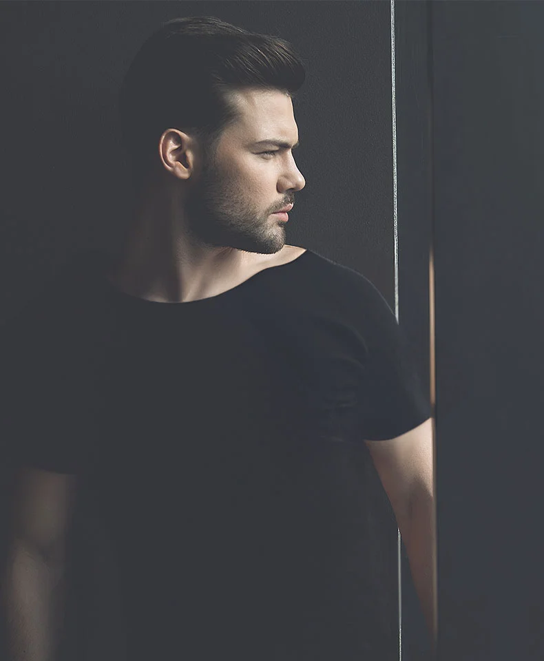Close-up profile of a light-skinned man with short, dark hair and a short beard. He gazes to his right with a neutral expression, wearing a fitted dark t-shirt. Soft lighting highlights his features against a dark gray background with subtle shadows. - Liporoscopic Hernia Repair in Coral Gables, FL