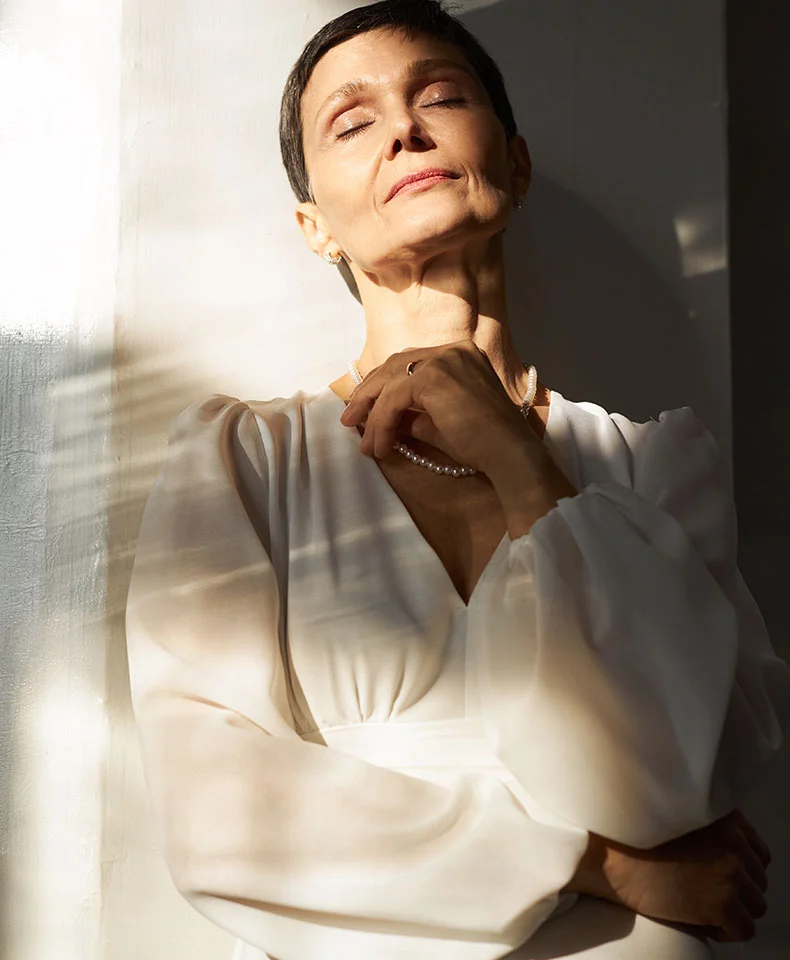 A woman with short, dark brown hair leans against a light wall, eyes closed in serenity. Soft window light highlights her loose, off-white blouse and delicate pearl-like necklace. The plain background enhances the tranquil mood. - Breast Lumpectomy in Coral Gables, FL