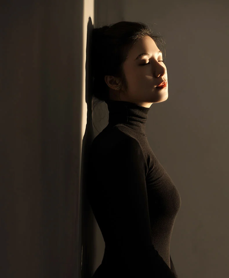 A woman in profile, leaning against a wall, has dark hair styled in a bun and eyes closed. She wears a dark-colored turtleneck top, with soft lighting casting gentle shadows on her face and shoulder. The neutral background and subtle contrasts focus attention on her relaxed, contemplative expression. - Breast Reduction Lift in Coral Gables, FL