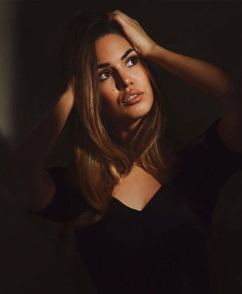 A woman with long, light brown hair gazes upwards, holding her hair near her forehead. She wears a black v-neck top, with dramatic lighting highlighting her face against a dark background. - Brow Lift in Coral Gables, FL