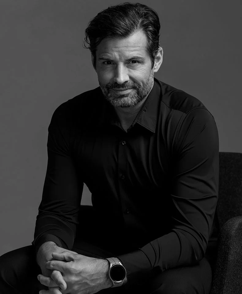 A black and white studio portrait of a man seated with his hands clasped in his lap. He wears a dark button-down shirt and a metallic wristwatch. His hair is dark and neatly combed, and he has a short, trimmed beard. The neutral gray background and focused lighting create a defined silhouette, highlighting his neutral, thoughtful expression. - Colonoscopy in Coral Gables, FL
