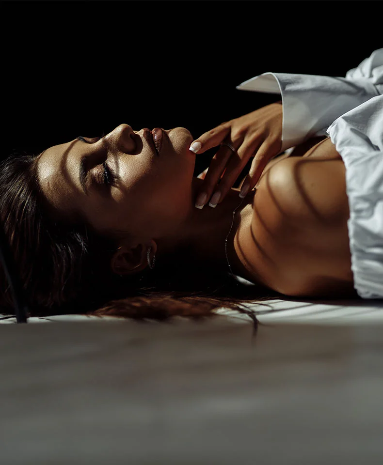 A woman lies on a bed, partially draped in a white blouse. Her dark hair spills over the sheets, and she has a serene expression. Soft lighting highlights her tanned skin, casting gentle shadows on her face and shoulders. - Dermal Fillers in Coral Gables, FL