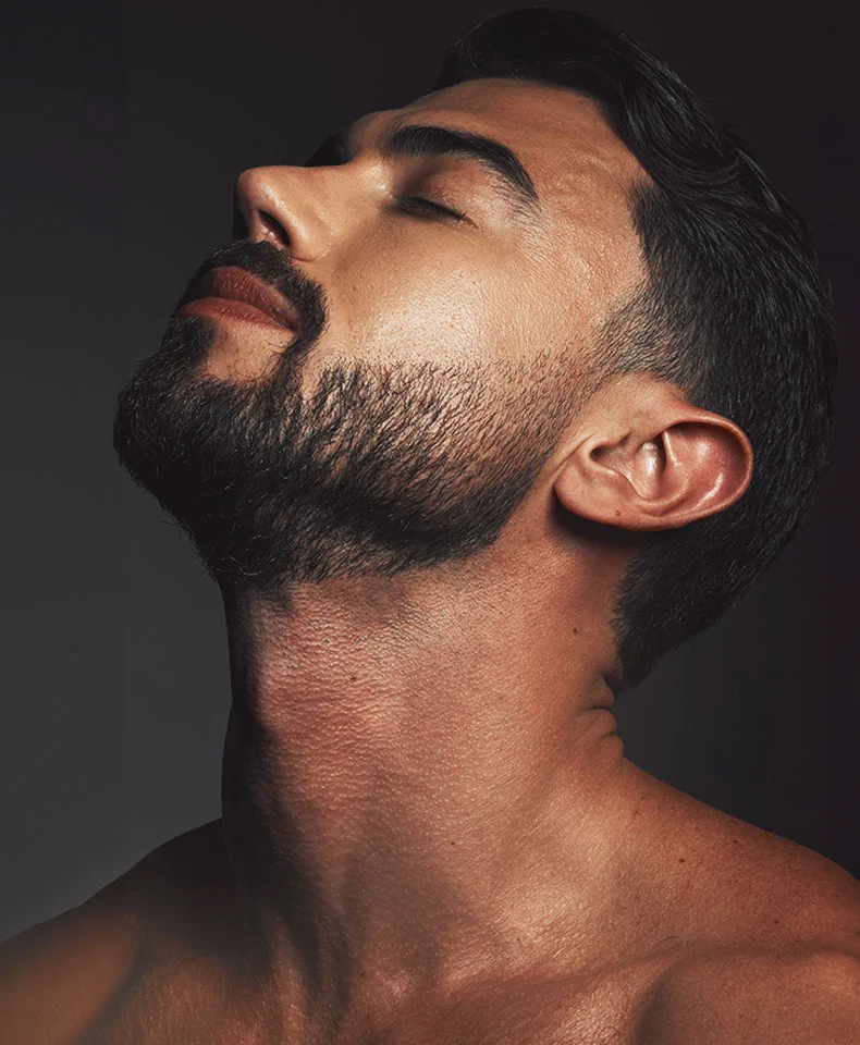 Close-up of a man’s head and upper torso with a relaxed, serene expression. His face is turned slightly to the right, with closed eyes, and he has a full, dark beard and mustache. His skin has a warm, light brown tone, and his dark hair is groomed. The dark gray-black background highlights the subject’s illuminated face and body, emphasizing the texture of his skin and beard. - EGD in Coral Gables, FL