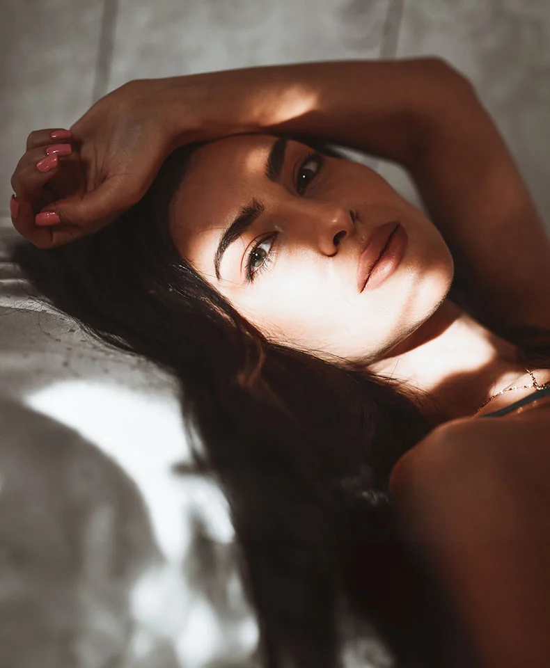 A young woman lies on a bed, her dark hair framing her face. She gazes serenely at the camera, with soft shadows highlighting her features. A light-colored necklace is visible, and the background is blurred. - Eyelid Surgery Blepharoplasty in Coral Gables, FL