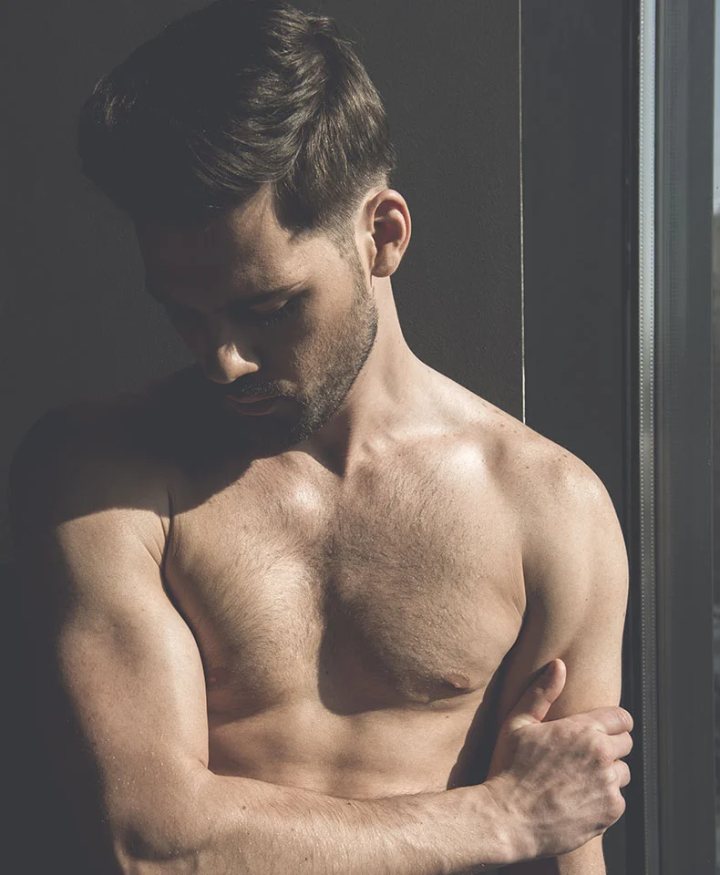 A shirtless man with a muscular build gazes downward, his short, dark hair styled neatly. The dark gray background features subtle lighting and shadows that emphasize his form, with a vertical seam or window frame adding depth. - F to M Breast Reconstruction in Coral Gables, FL