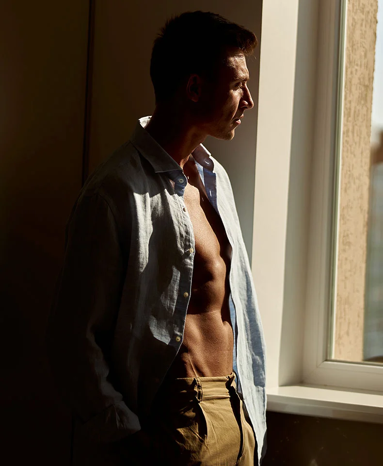 A man stands in profile, gazing out a window. He has short dark hair, a tan complexion, and a muscular build. He wears a light-blue linen shirt open over his toned torso, paired with light khaki pants. The beige background features strong shadows, suggesting light from the right. The focus is on the man’s physique and the window, with the surrounding environment blurred. - Gynecomastia in Coral Gables, FL