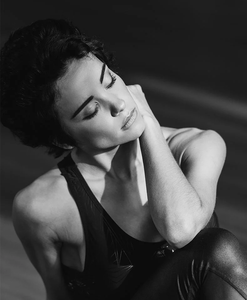 Close-up black and white portrait of a woman with serene expression, head down, and eyes closed. She wears a dark satin-like leotard, with soft lighting highlighting her form against a dark backdrop. - Laser Skin Resurfacing in Coral Gables, FL