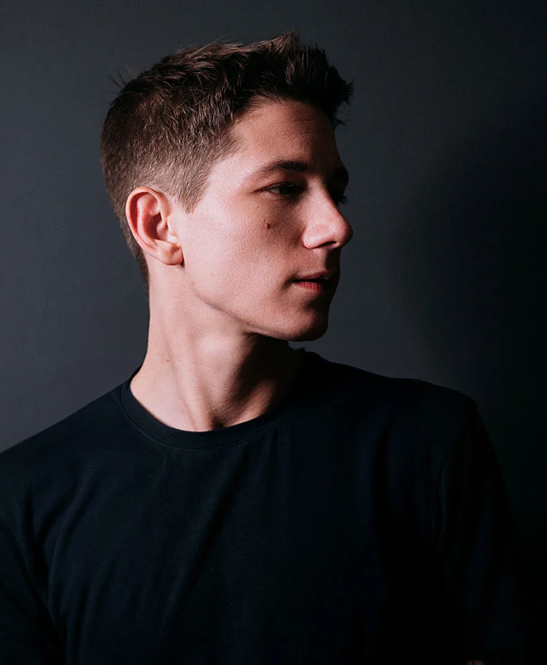 A close-up of a young man in profile, with short textured brown hair and a light complexion. He wears a black t-shirt, and the lighting highlights his face and upper body, with subtle shadows. The dark gray background keeps the focus on him, creating a professional feel. - Mole Removal in Coral Gables, FL