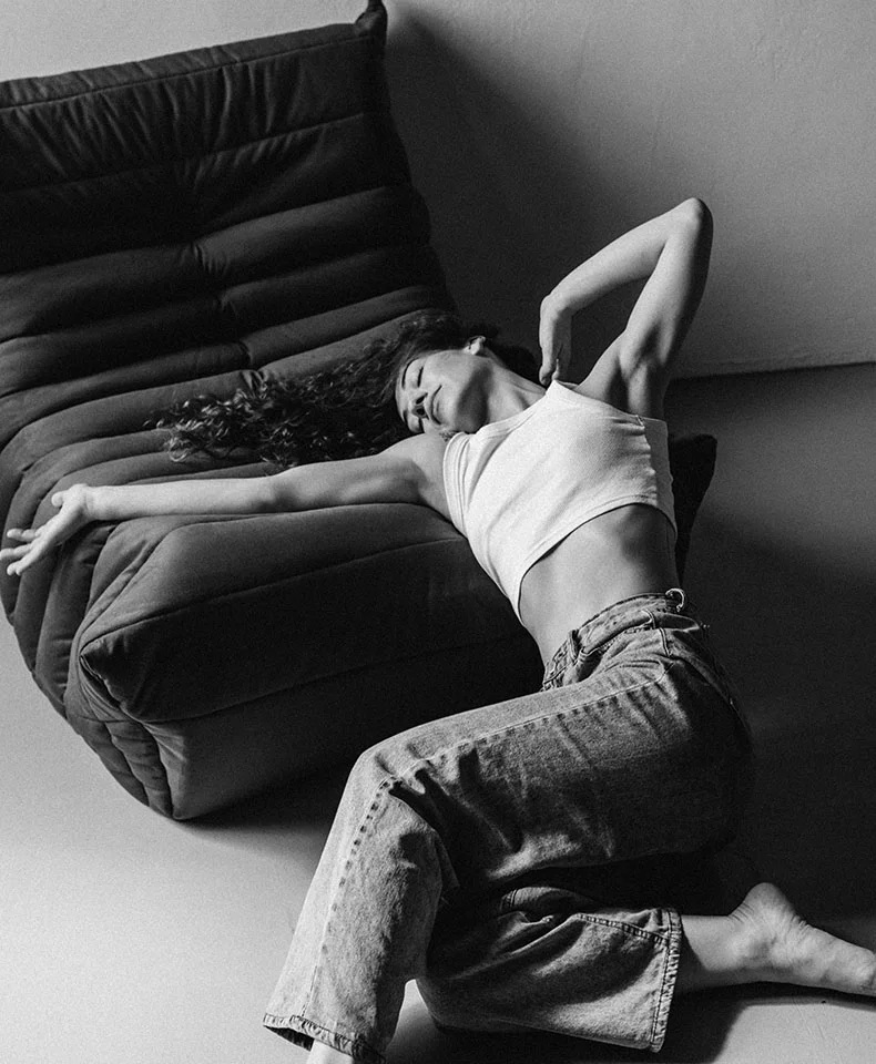 A black and white full shot of a woman lounging diagonally on a dark modular couch, wearing a cropped white tank top and light wash denim jeans. Her dark curly hair frames her face as she gazes slightly upwards. The quilted couch design and soft directional lighting highlight her relaxed pose against a plain background. - Scarr Removal in Coral Gables, FL