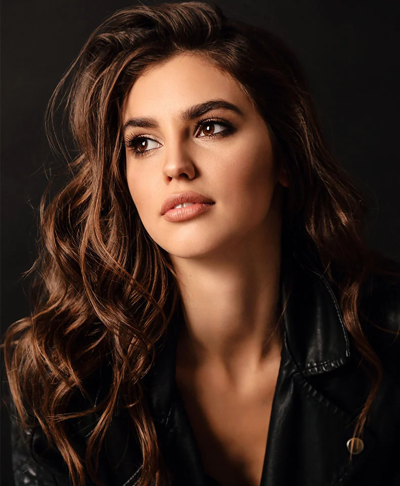 A close-up portrait of a young woman with shoulder-length wavy brown hair, subtle makeup, and a pleasant expression looking off to the side. She wears a black leather jacket, and the dark background highlights her features. Soft lighting focuses on her face and upper body, emphasizing her natural beauty and style. - Skinvive in Coral Gables, FL