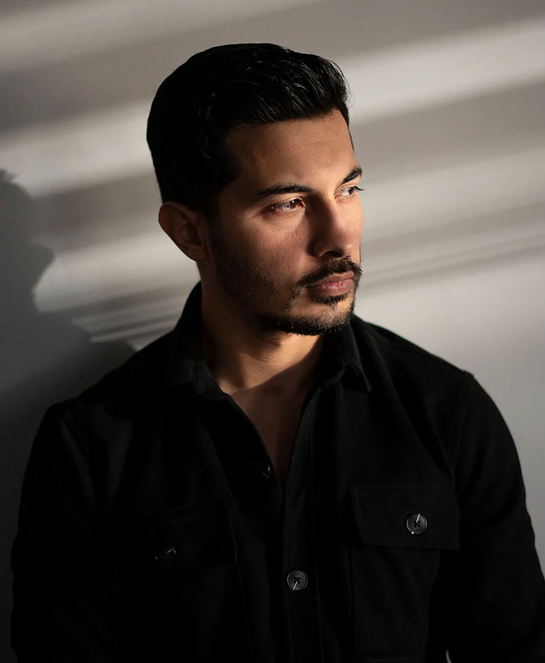 A close-up of a medium-built man with short, dark brown hair, looking slightly to his right with a thoughtful expression. He has a short, neatly trimmed beard and mustache and is wearing a black button-down shirt or jacket, possibly a wool blend. The light gray or beige wall behind him has subtle shadows, adding depth and mood to the image. - Soft Tissue Biopsy in Coral Gables, FL