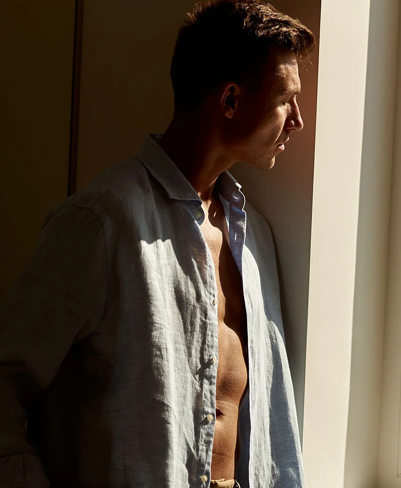 A mid-chest-up profile shot of a man with short, dark brown hair and a light tan complexion. He wears a light grayish-blue linen shirt, unbuttoned from the mid-chest up, revealing his toned torso. Natural sunlight casts shadows, emphasizing the shirt’s texture and his form. A window in the background provides soft illumination, with a light beige or cream wall on the opposite side. - Ventral Hernia Repair in Coral Gables, FL
